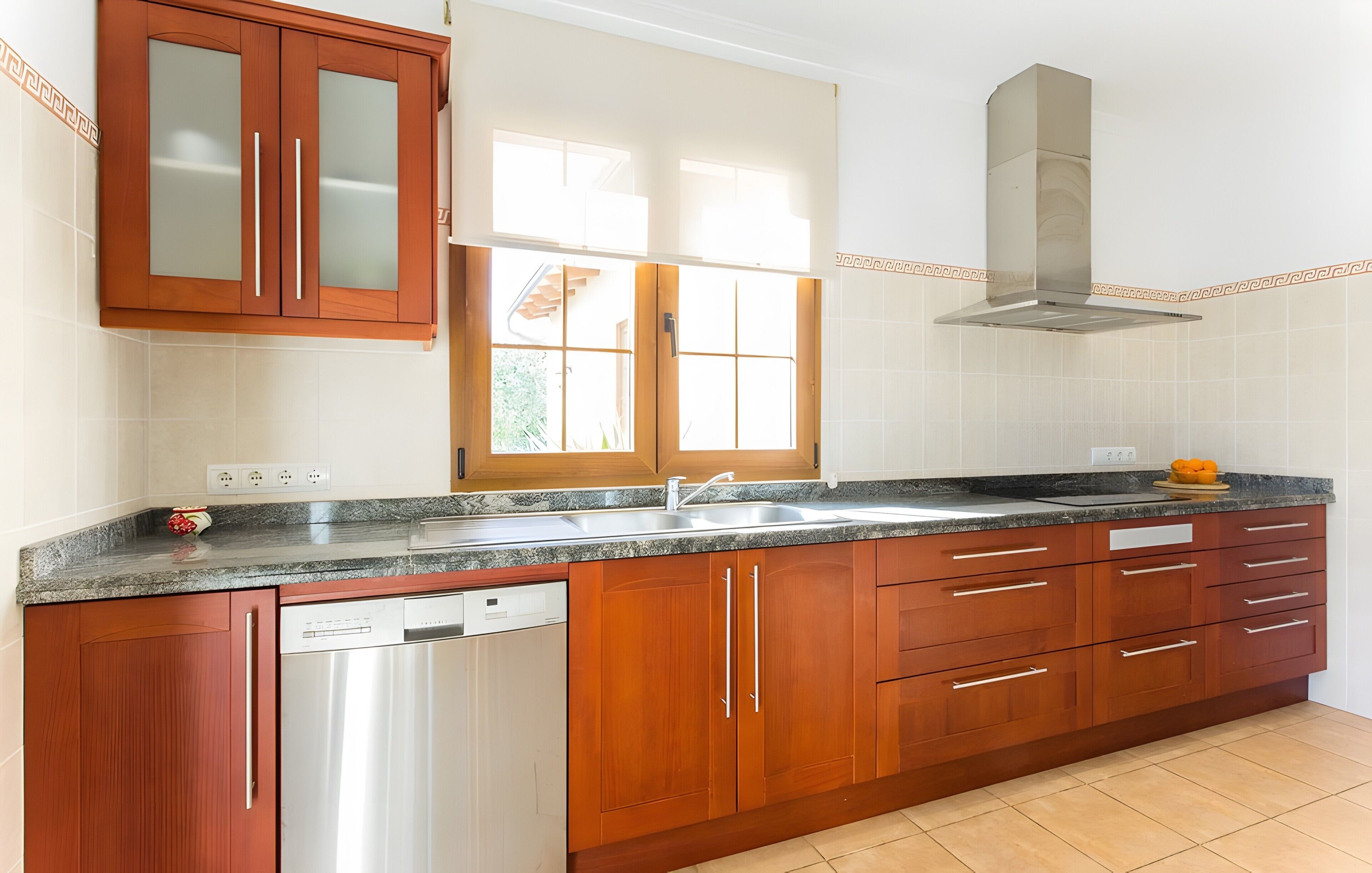 Una cocina con armarios de madera, lavavajillas de acero inoxidable, encimera de granito, una ventana y una campana extractora de acero inoxidable.