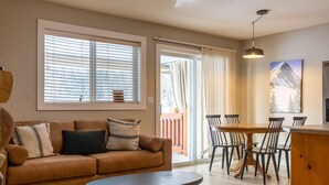 A modern living room with a sofa, a dining table with chairs, and a large window with blinds. - Newly Renovated 3 Bedroom l A/C | Sleeps 8 (Fernie)