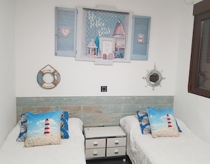 Un dormitorio con dos camas, una estantería con objetos decorativos, una mesita y una ventana con contraventanas.
