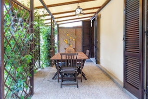 A covered outdoor seating area with a table and chairs, a decorative vase with flowers, and a lattice fence.