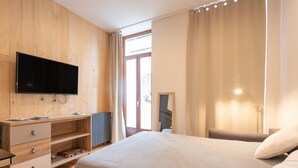 A modern bedroom with a flat-screen TV, wooden panel walls, a bed with a grey bedspread, a bedside table, and a chair.