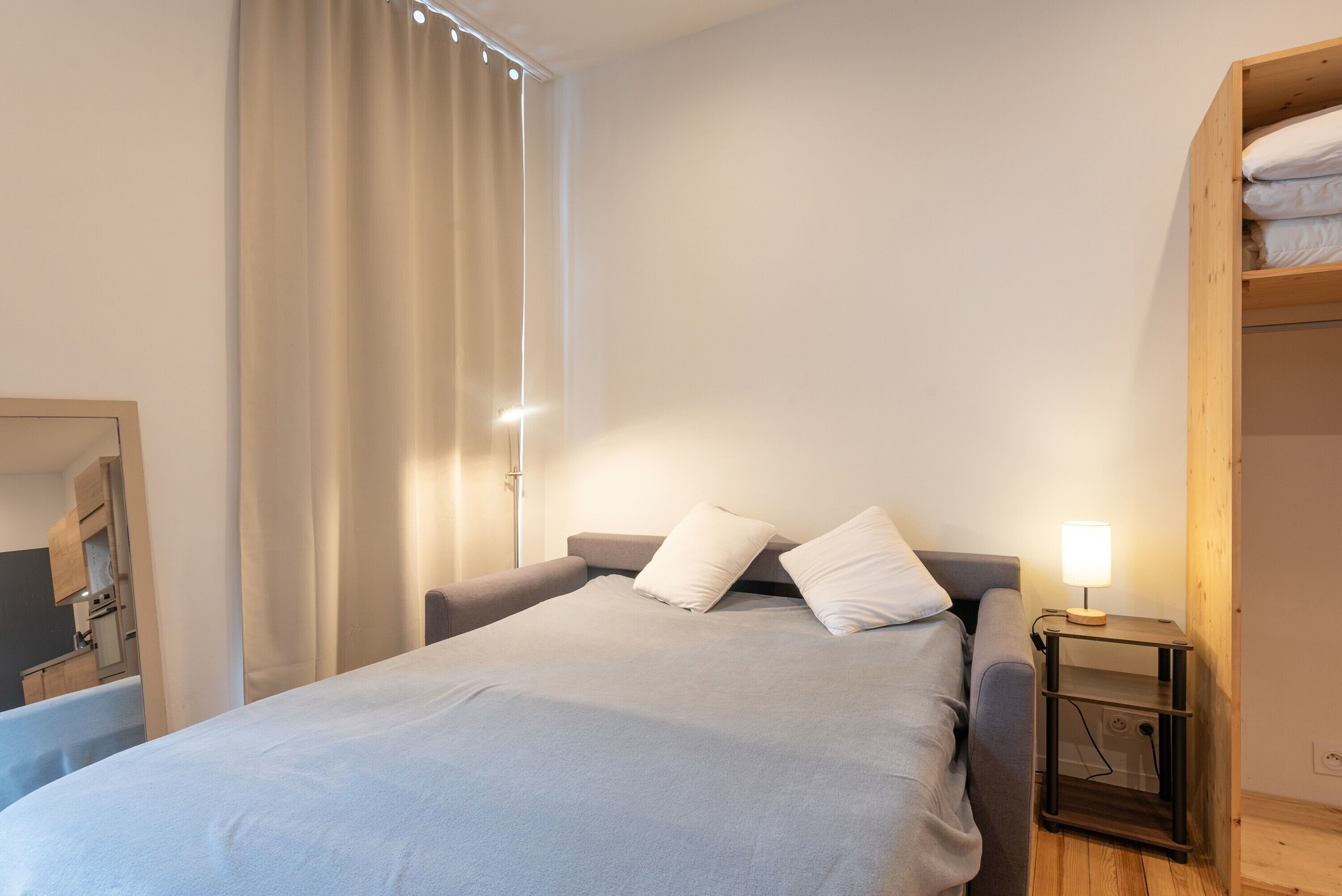 Ein Schlafzimmer mit einem Bett, einem Nachttisch, einem Holzschrank, einem Fenster mit Vorhängen und einem Spiegel.