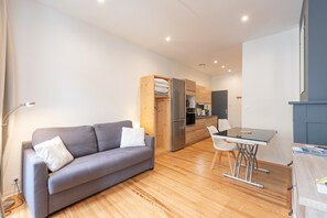 Un salon moderne avec un canapé gris, un meuble en bois, une table à manger avec des chaises, et un coin cuisine à l’arrière-plan.