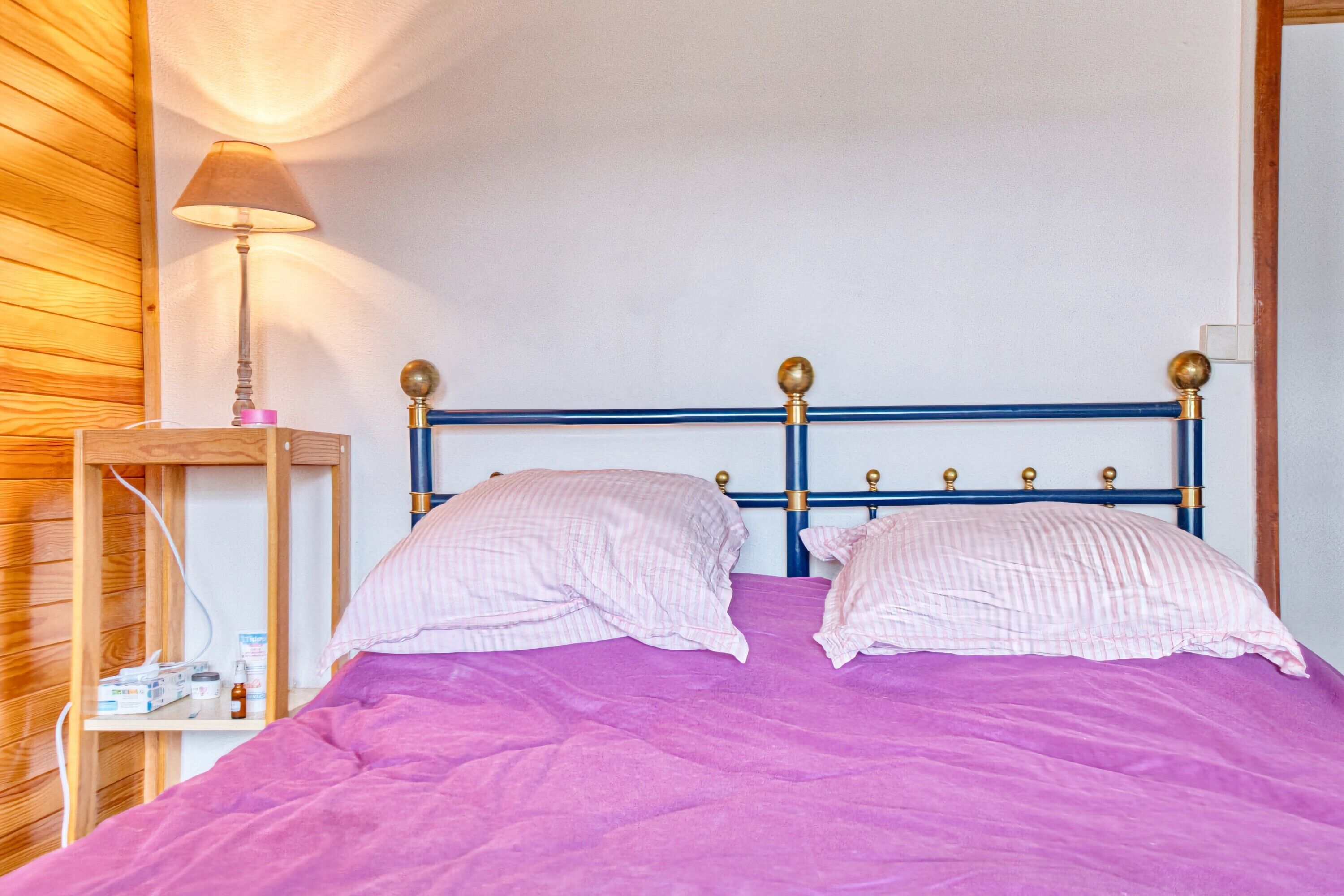 Ein Bett mit violetter Bettdecke, zwei Kissen, ein Nachttisch aus Holz mit Lampe und ein blaues Kopfteil mit goldfarbenen Akzenten.