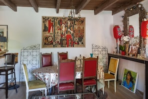 A dining area with a table set for four, surrounded by chairs with red upholstery. A large, colorful painting hangs above the table, and a decorative mirror with red lanterns is on the wall to the right.