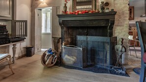 Ein Kamin mit Kaminfeuer, verziert mit roten Blumen, einem Spiegel und einem Sonnendekor. Neben dem Kamin steht ein Holzkorb, und im Vordergrund ist ein Stuhl zu sehen.