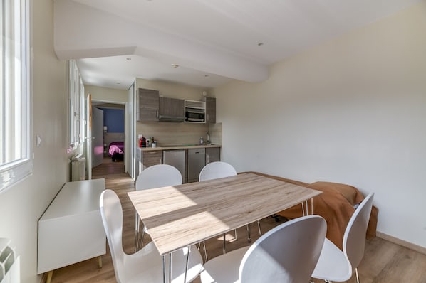 Una cocina moderna con electrodomésticos integrados, una zona de comedor con mesa de madera y sillas blancas, y vistas a un dormitorio.