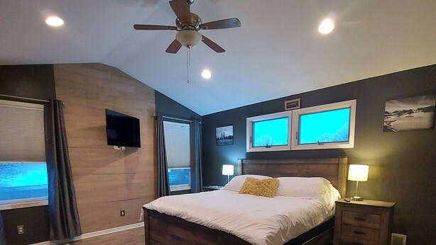 A bedroom with a bed, a ceiling fan, two wall-mounted TVs, a bedside table with a lamp, and a window with curtains.