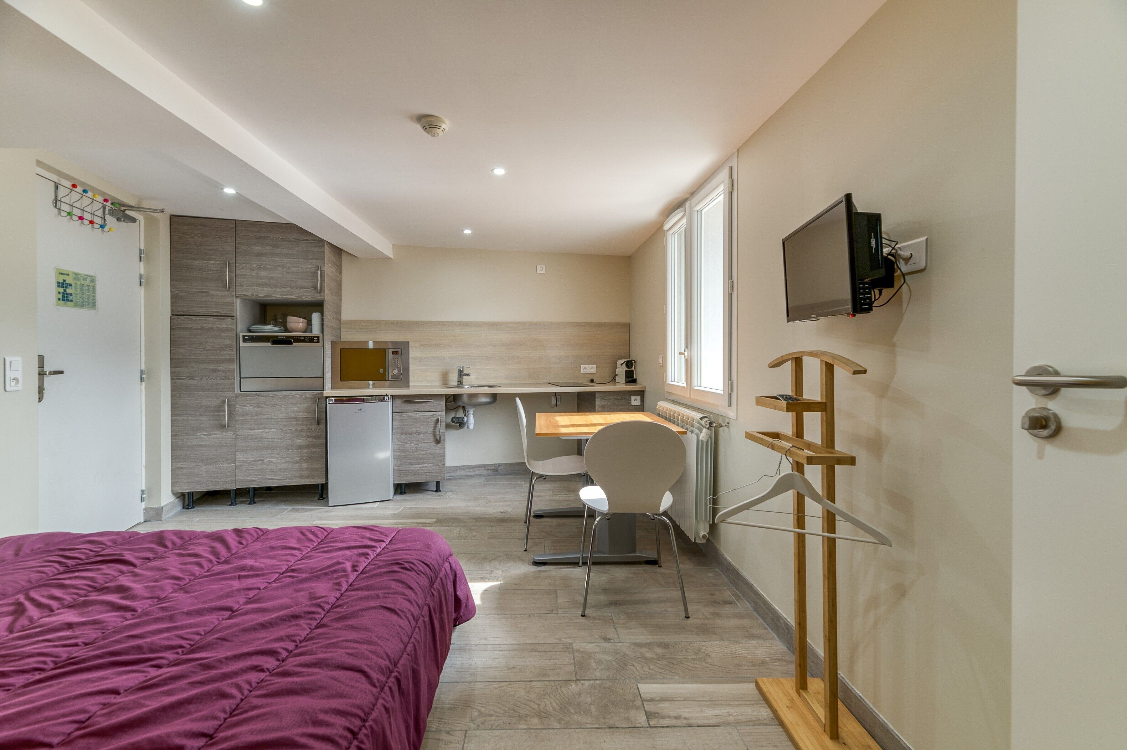 Ein kleines Hotelzimmer mit Bett, Küchenzeile, Essbereich und an der Wand befestigtem Fernseher.