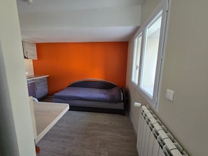 A compact room with a bed, a small kitchenette, and a window providing natural light.