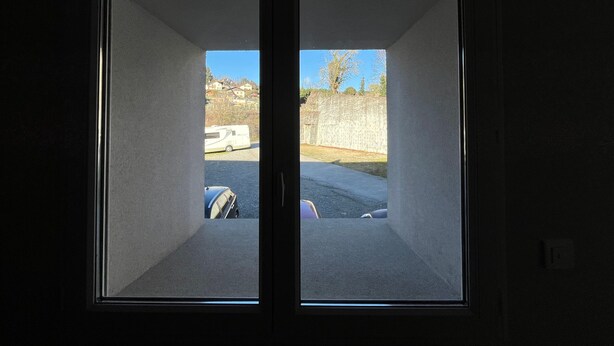 Ein Zimmer mit großem Fenster, einer Tür und einem sichtbaren Außenbereich mit geparktem Fahrzeug und einem Gebäude im Hintergrund.