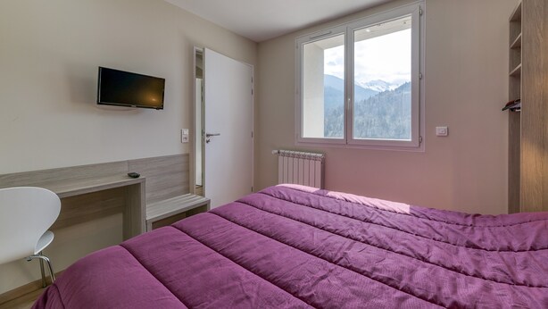 Een slaapkamer met een bed, een aan de muur bevestigde televisie, een raam met uitzicht op bergen en een radiator.