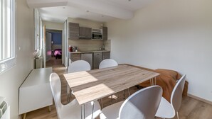 Een moderne keuken met ingebouwde apparatuur, een eethoek met een houten tafel en witte stoelen, en uitzicht op een slaapkamer.