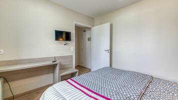 A bedroom with a bed, a bedside table, a TV mounted on the wall, and a door leading to another room.