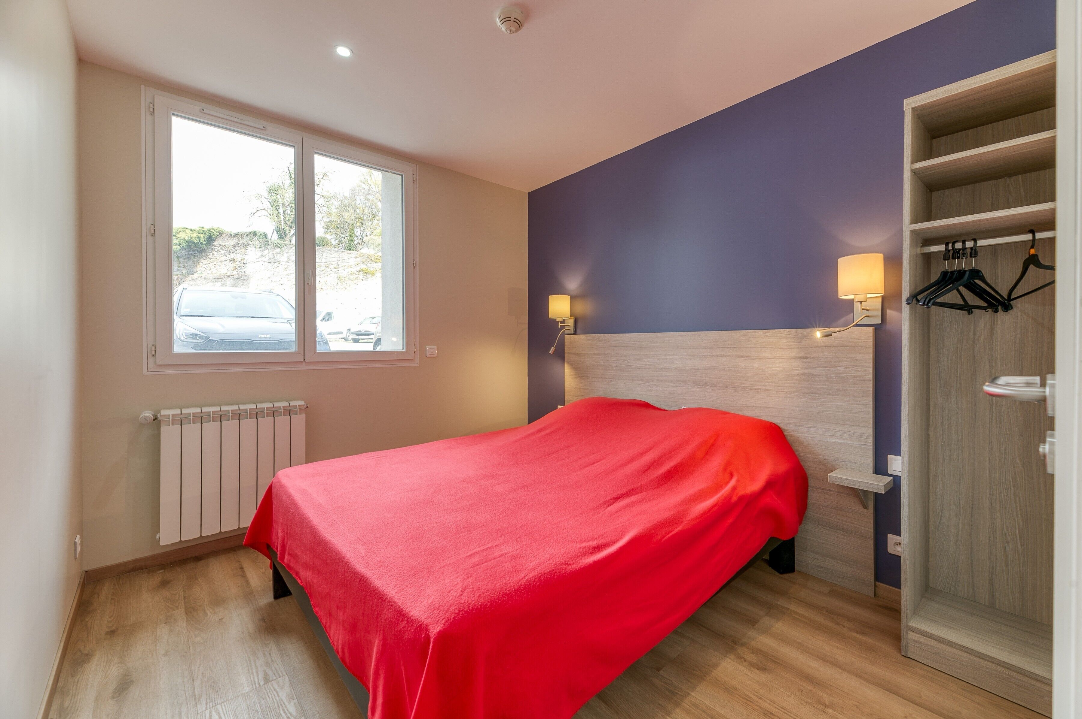 A bedroom with a bed, a window, a radiator, a wall-mounted light, and a wardrobe.