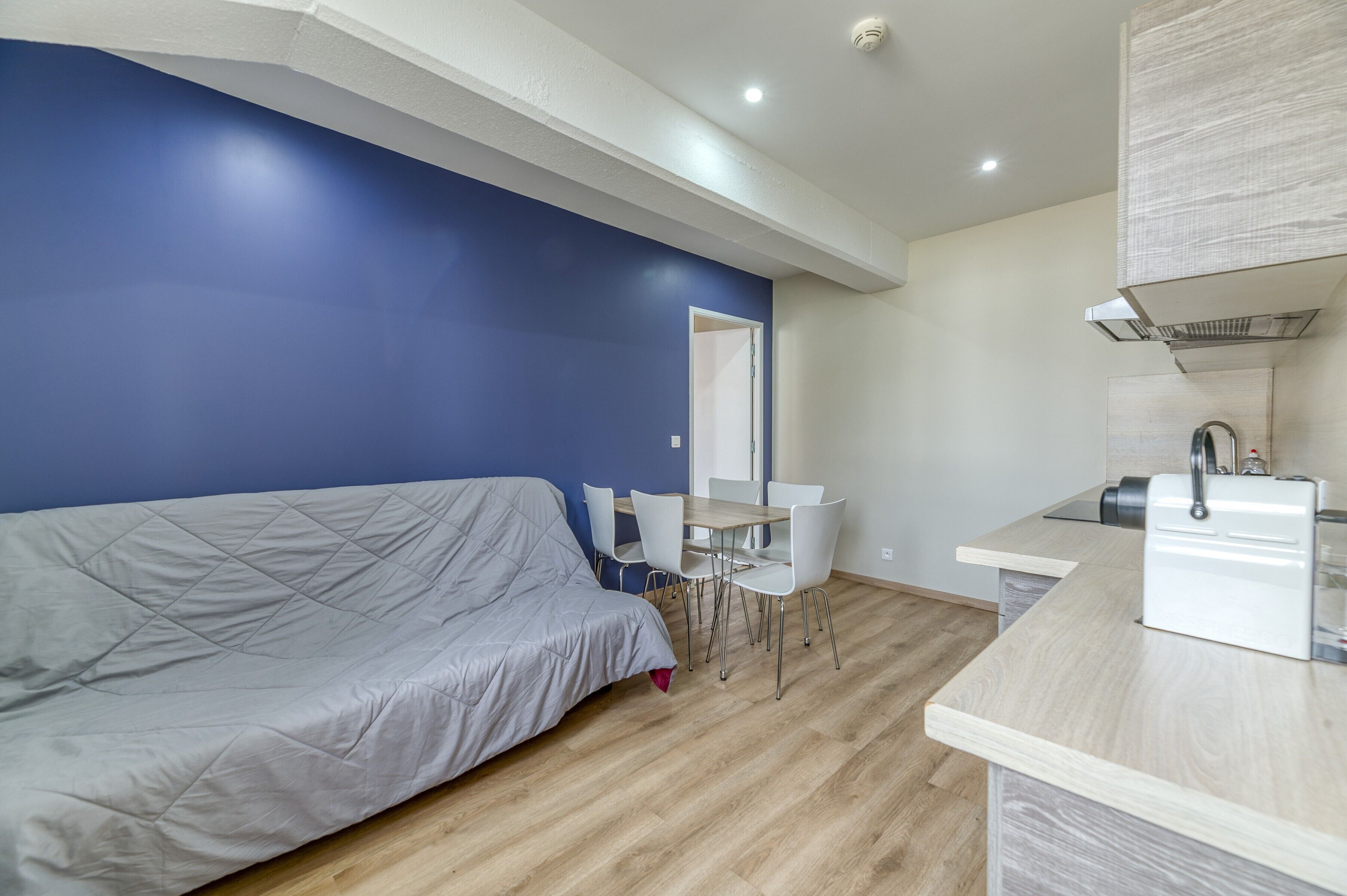 A modern room with a bed, a dining table with chairs, a kitchen area, and a blue accent wall.