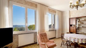 Quarto com televisão, uma cadeira de balanço, mesa de jantar com cadeiras e vista através de janelas amplas.