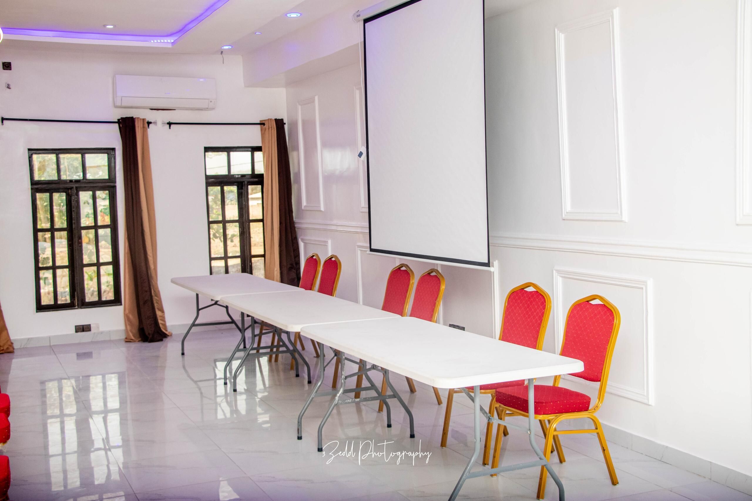A conference room with a long table, red chairs, a projector screen, and windows with curtains.