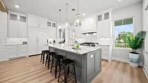 Una cocina moderna con armarios blancos, una isla grande con taburetes, iluminación colgante y vistas al exterior.