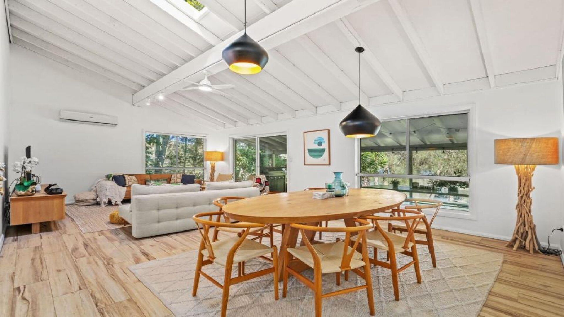 A spacious living area with a wooden dining table, white sofas, and large windows offering a view of greenery.