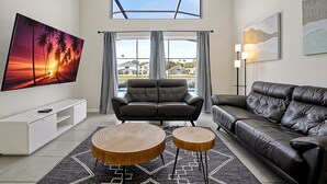 Ein modernes Wohnzimmer mit großem Fernseher, zwei Sofas, einem Couchtisch und Blick auf Häuser durch große Fenster.