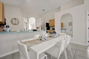 A dining area with a white table and chairs, a clock on the wall, and a kitchen in the background. - Magical Disney Retreat | Lake Berkley Resort & Pool (Kissimmee)