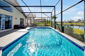 An indoor swimming pool with a glass enclosure, a seating area with a sofa and chairs, and a view of a lake and buildings in the background. - Magical Disney Retreat | Lake Berkley Resort & Pool (Kissimmee)