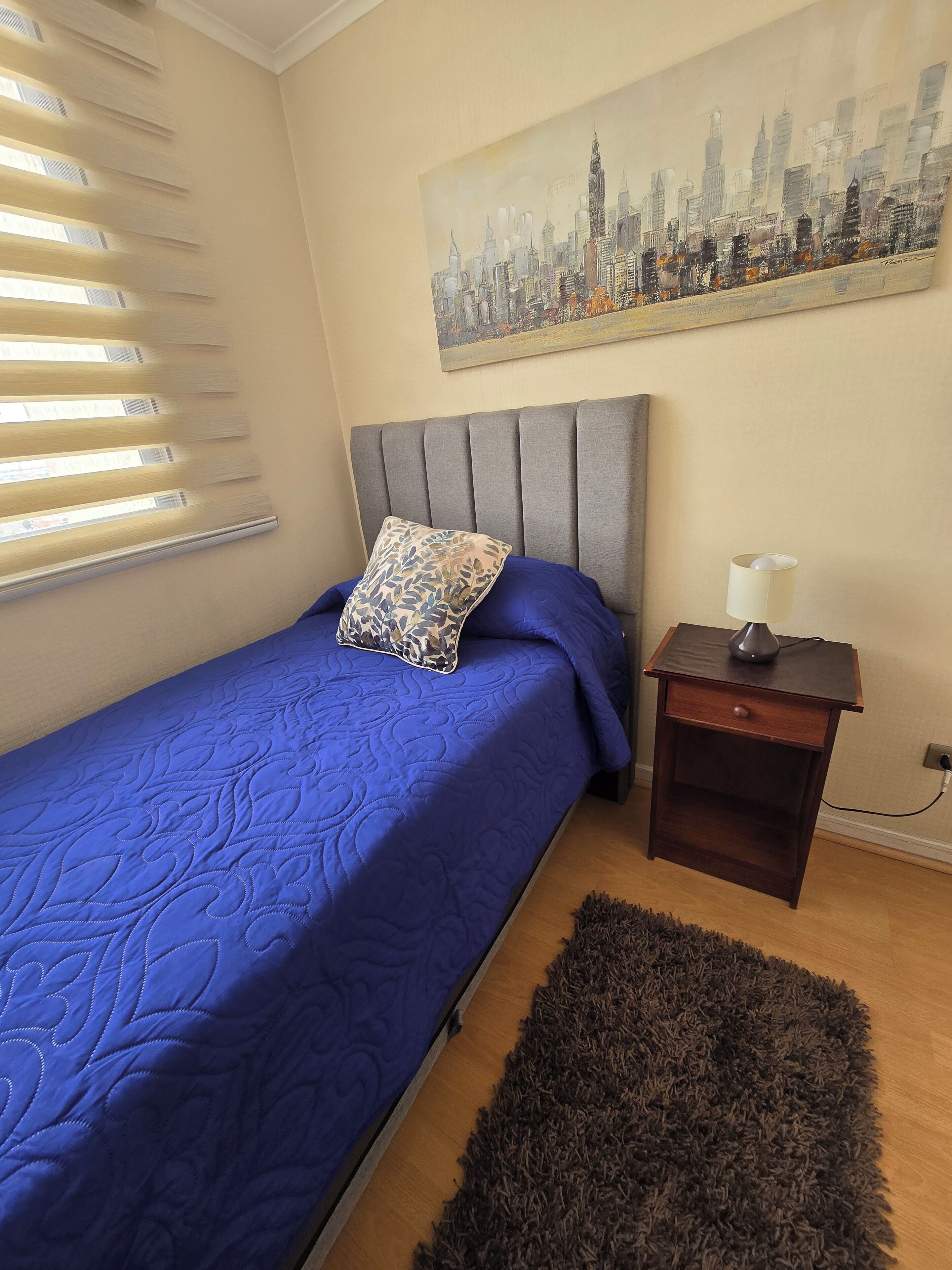 A bedroom with a bed, a nightstand, a lamp, a cityscape painting, and a window with blinds.