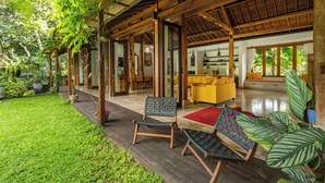 Una terraza de madera con dos sillas, un tapete rojo y un sofá amarillo en un espacio interior bien iluminado con ventanas grandes y vistas al verdor exterior.