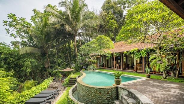 Una piscina rodeada de exuberante vegetación, una terraza de madera y un edificio con tejado de tejas.