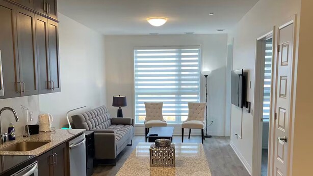 A modern kitchen with granite countertops, stainless steel appliances, and a central island. There is a dining area with a sofa and chairs, and a large window with blinds. - Executive Luxury Apartment. (Oakville)