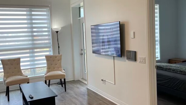A modern living room with a flat-screen TV mounted on the wall, two armchairs, a coffee table, and a large window with blinds. - Executive Luxury Apartment. (Oakville)
