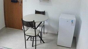 Een kleine kamer met een witte tafel en twee stoelen, een witte koelkast, een houten deur en een tv aan de muur.