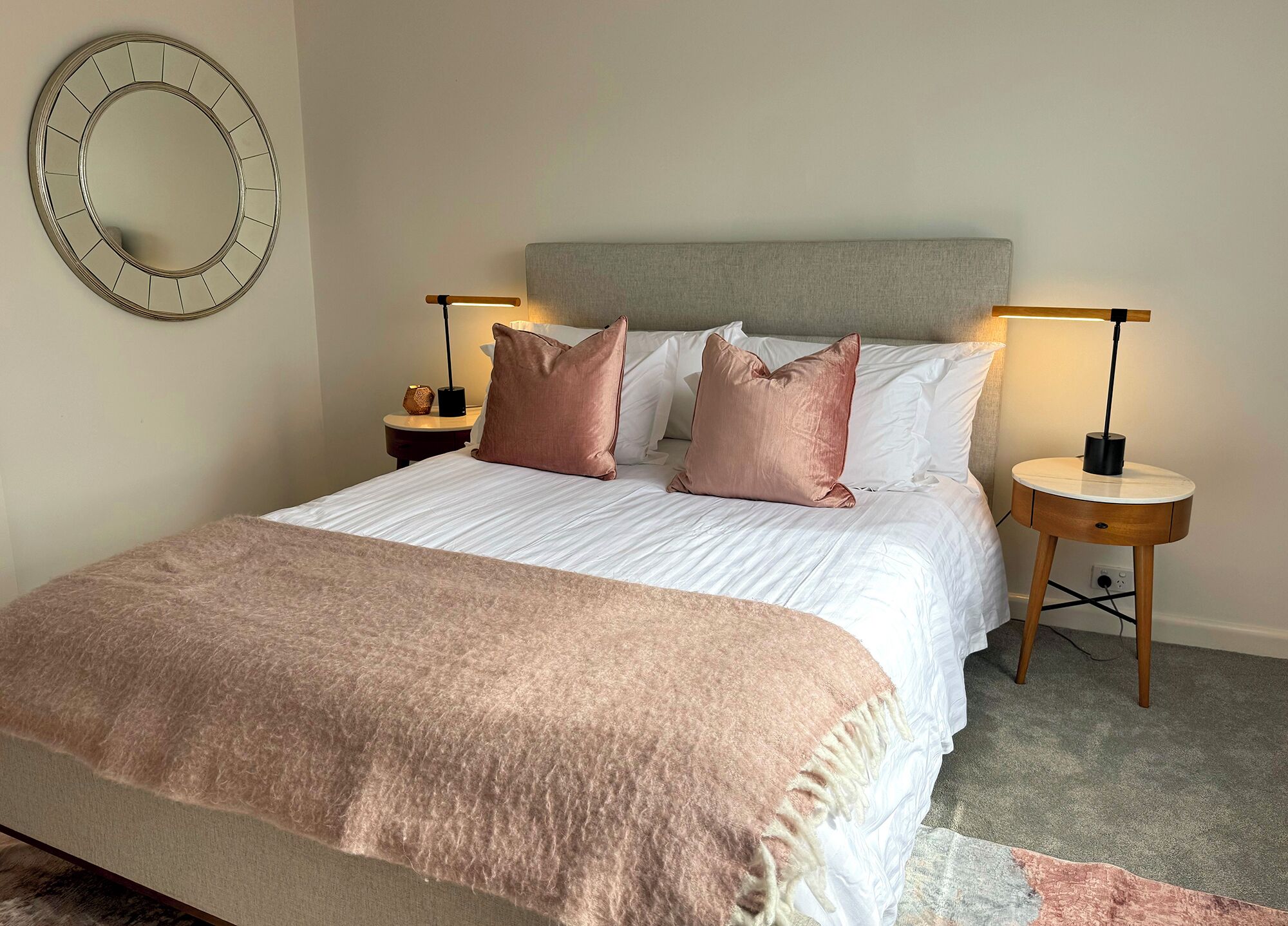 A neatly made bed with a grey headboard, two bedside tables with lamps, and a round mirror on the wall.