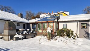 A snow-covered house with a stone fireplace, a glass-enclosed porch, and a yellow building in the background. - Nice home in Fohnsdorf with sauna (Fohnsdorf)