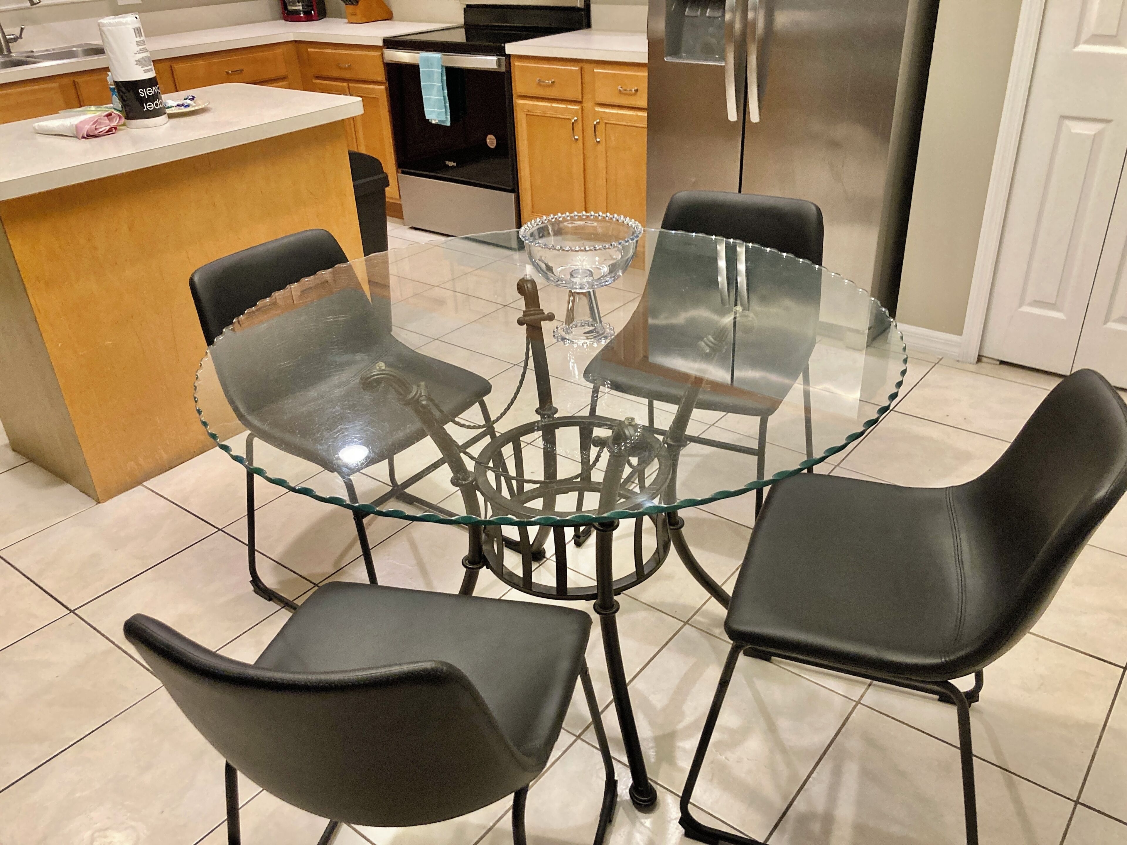 A modern kitchen with a round glass table surrounded by four black chairs, a stainless steel refrigerator, and wooden cabinets.