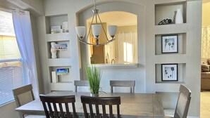 A dining area with a wooden table and chairs, a chandelier, and shelves with framed pictures and decorative items.