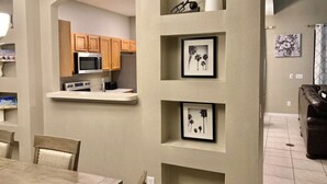 A modern kitchen with wooden cabinets, a microwave, and a countertop. There are framed pictures on the wall and a dining area with chairs.