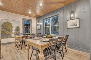 A dining area with a wooden table and chairs, framed artwork on the walls, and large windows.