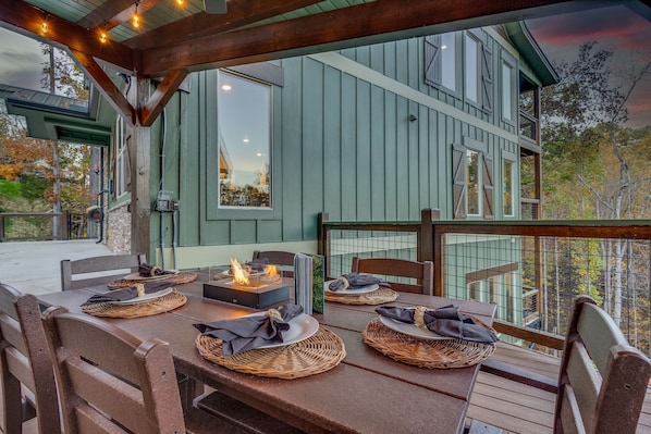 A dining area with a fire feature, wooden furniture, and a view of a green building.