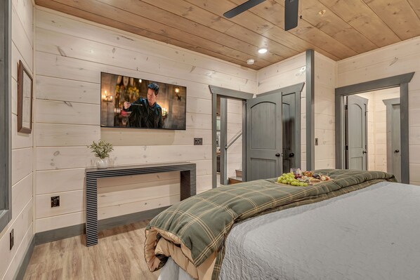 A bedroom with a bed, a TV mounted on the wall, a fireplace, and a door leading to another room.