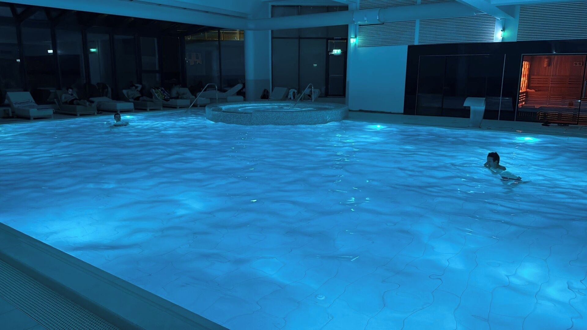 Ein großer, überdachter Pool mit einem zentralen Whirlpool, Liegestühlen und einem durch Glas geschützten Bereich.