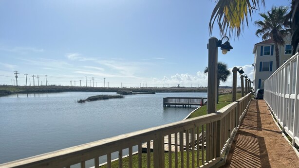 Um deck de madeira com guarda-corpo, com vista para um corpo d'água calmo, uma área gramada e um edifício com fachada branca ao fundo.