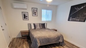 Een slaapkamer met een bed, houten vloeren, een nachtkastje, een dressoir, een raam met jaloezieën en een lijst met een afbeelding aan de muur.