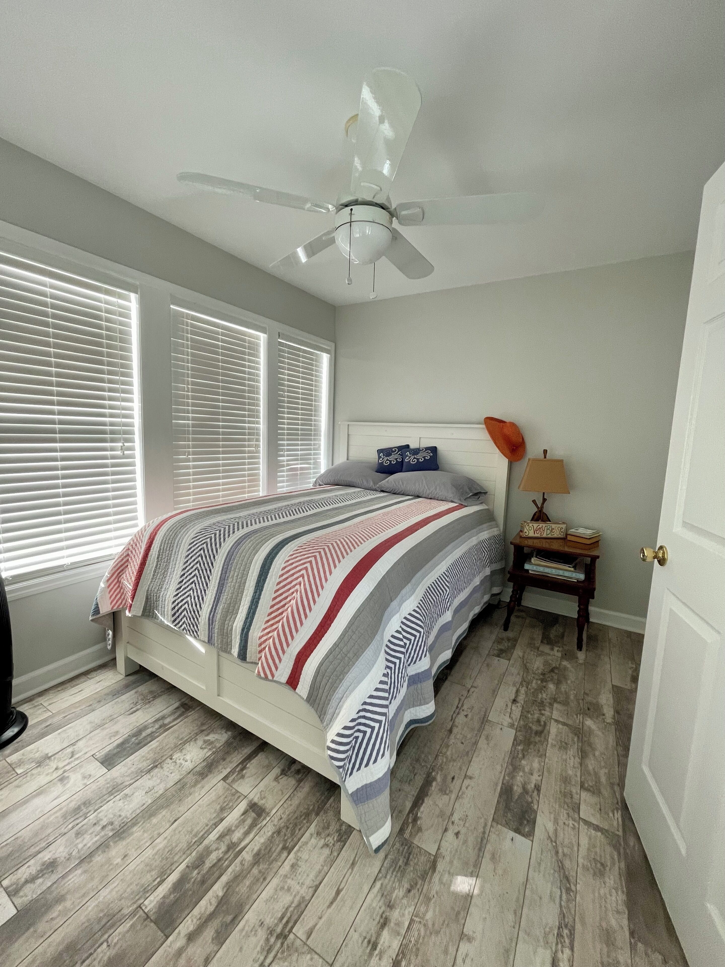 Quarto com cama, ventoinha no teto, mesa de cabeceira com abajur e uma porta.