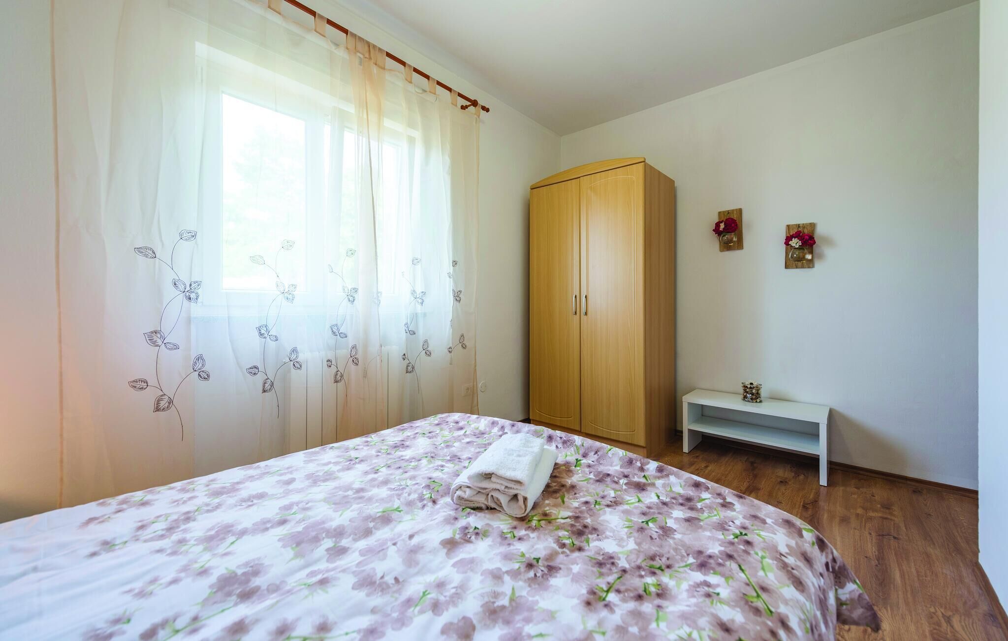 Quarto com cama, guarda-roupa de madeira, pequeno banco branco e dois vasos de flores fixados na parede.