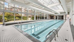 A modern indoor swimming pool with clear glass ceilings, a tiled floor, and a view of residential buildings outside.