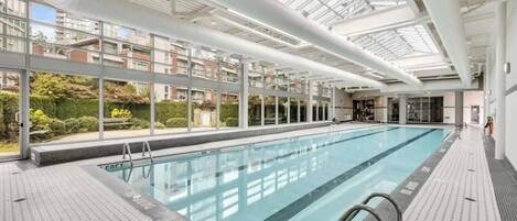 Una piscina interior moderna con techos de vidrio transparente, suelo de baldosas y vistas a edificios residenciales.