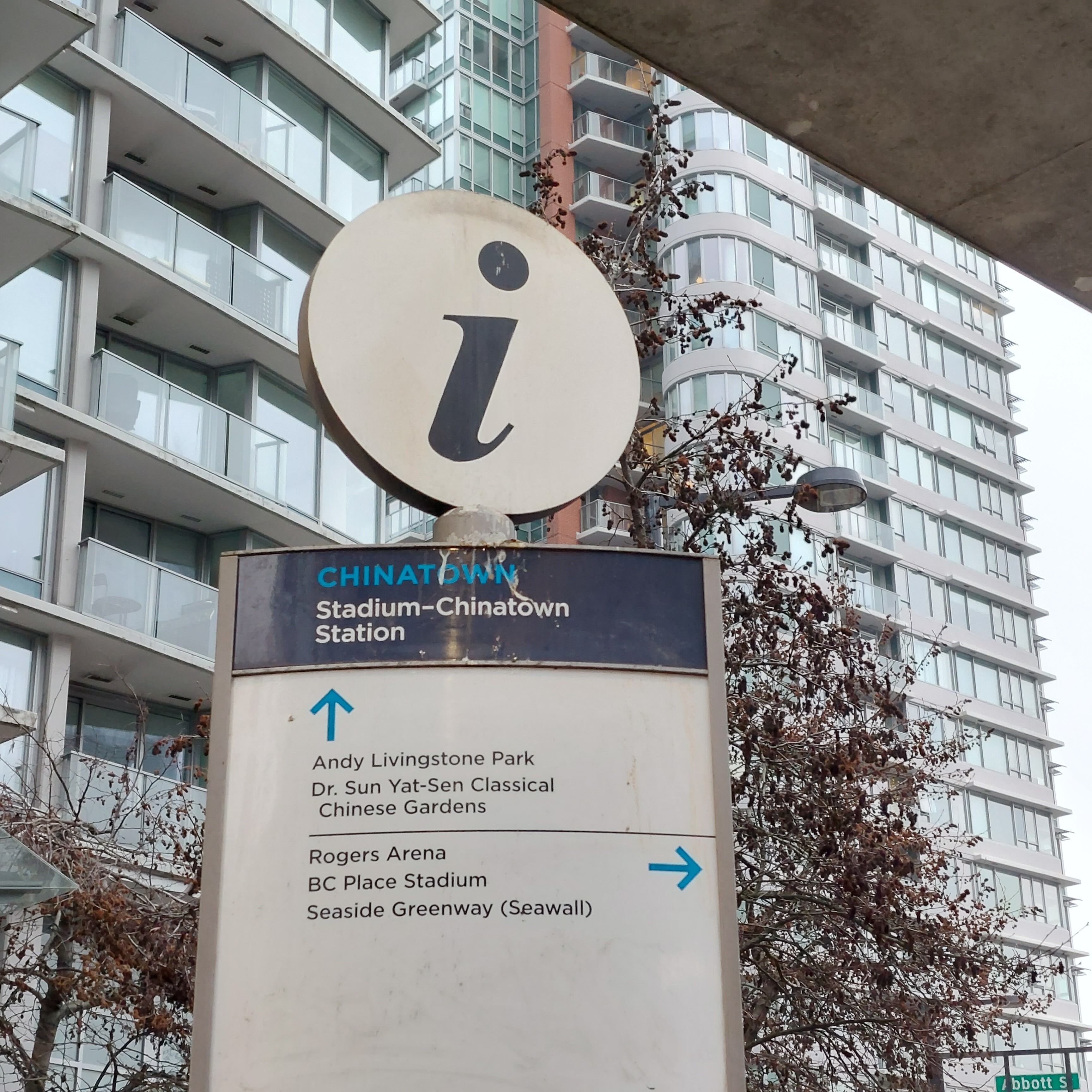 En vejskilte med et 'i'-symbol, der angiver informationsservice, der peger mod forskellige steder som Andy Livingston Park, Dr. Sun Yat-Sen Classical Chinese Gardens, Rogers Arena, BC Place Stadium og Seaside Greenway.
