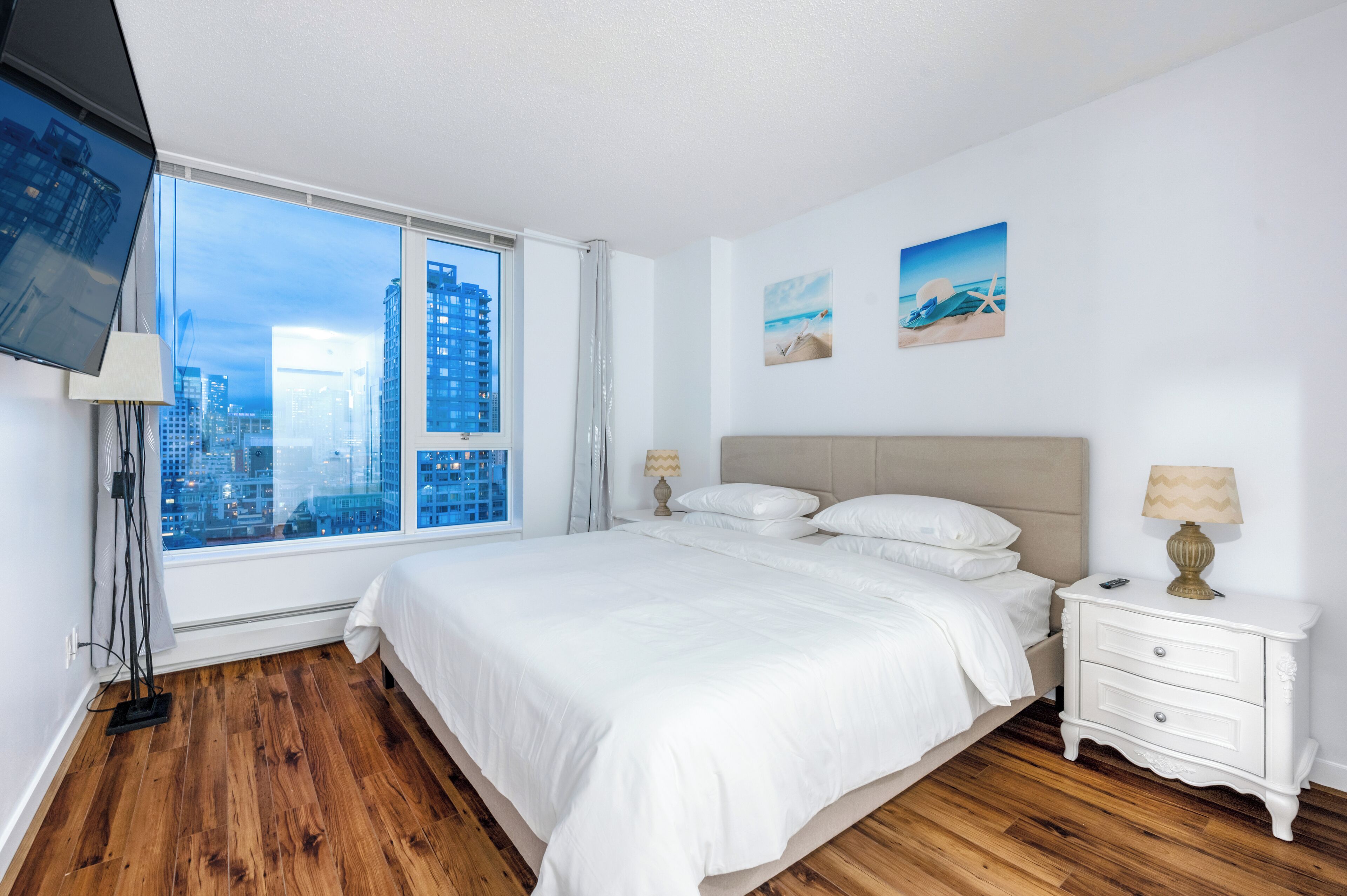 A modern hotel room with a large bed, a bedside table, a TV mounted on the wall, and a window offering a city view.
